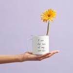 Hand holding a mug with a sunflower and motivational quote, set against a purple background.